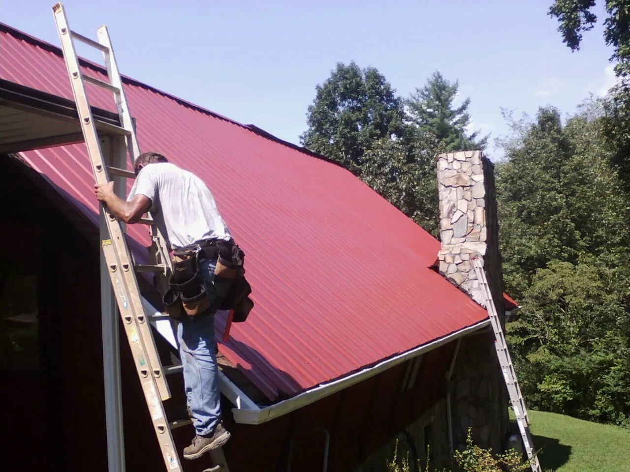 Roofer on a ladder beside a red metal roof and stone chimney — Soffit & Fascia Repair by Roofing Brothers in Van Buren