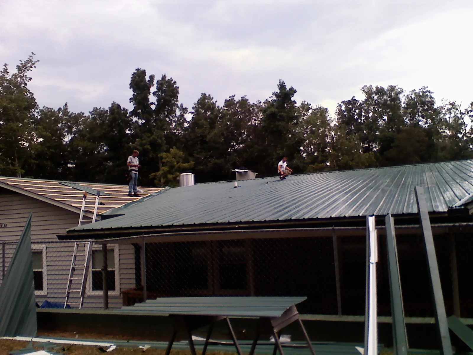 Two roofers installing green metal panels above a covered porch in Van Buren
